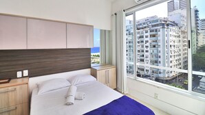 A hotel room with a large bed, a wooden headboard, a nightstand, a window with a city view, and a view of tall buildings. - Rio Spot Homes C056 (Rio de Janeiro)