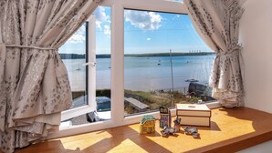 Una habitación con un gran ventanal con vista a una masa de agua, una mesa de madera con una maqueta de una casa y un libro, y cortinas con estampado floral.