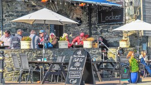 Un restaurant en bord de mer avec une terrasse, des murs en pierre et un tableau affichant le menu, où l’on peut lire les mentions « Davy Jones Locker » et « Café unique et charmant ».