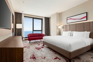 A hotel room with a large bed, a red armchair, a desk, and a view of the sea.