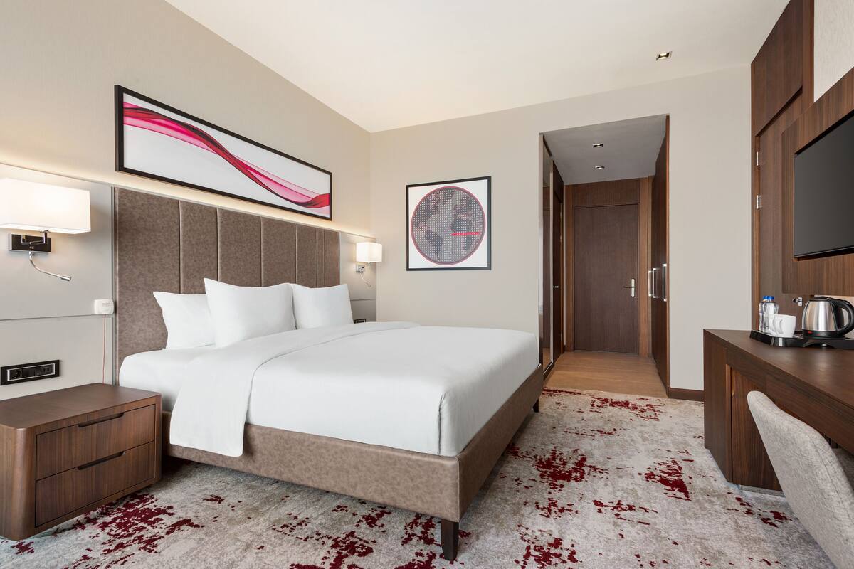 Ein Hotelzimmer mit einem großen Bett, einem Nachttisch, einem Schreibtisch mit Stuhl, einem Fernseher und Bildern an den Wänden.