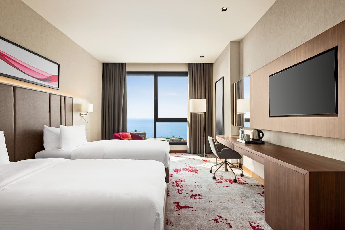 Ein Hotelzimmer mit zwei Betten, einem Schreibtisch, einem Stuhl, einem Fernseher und einem großen Fenster mit Blick aufs Meer.