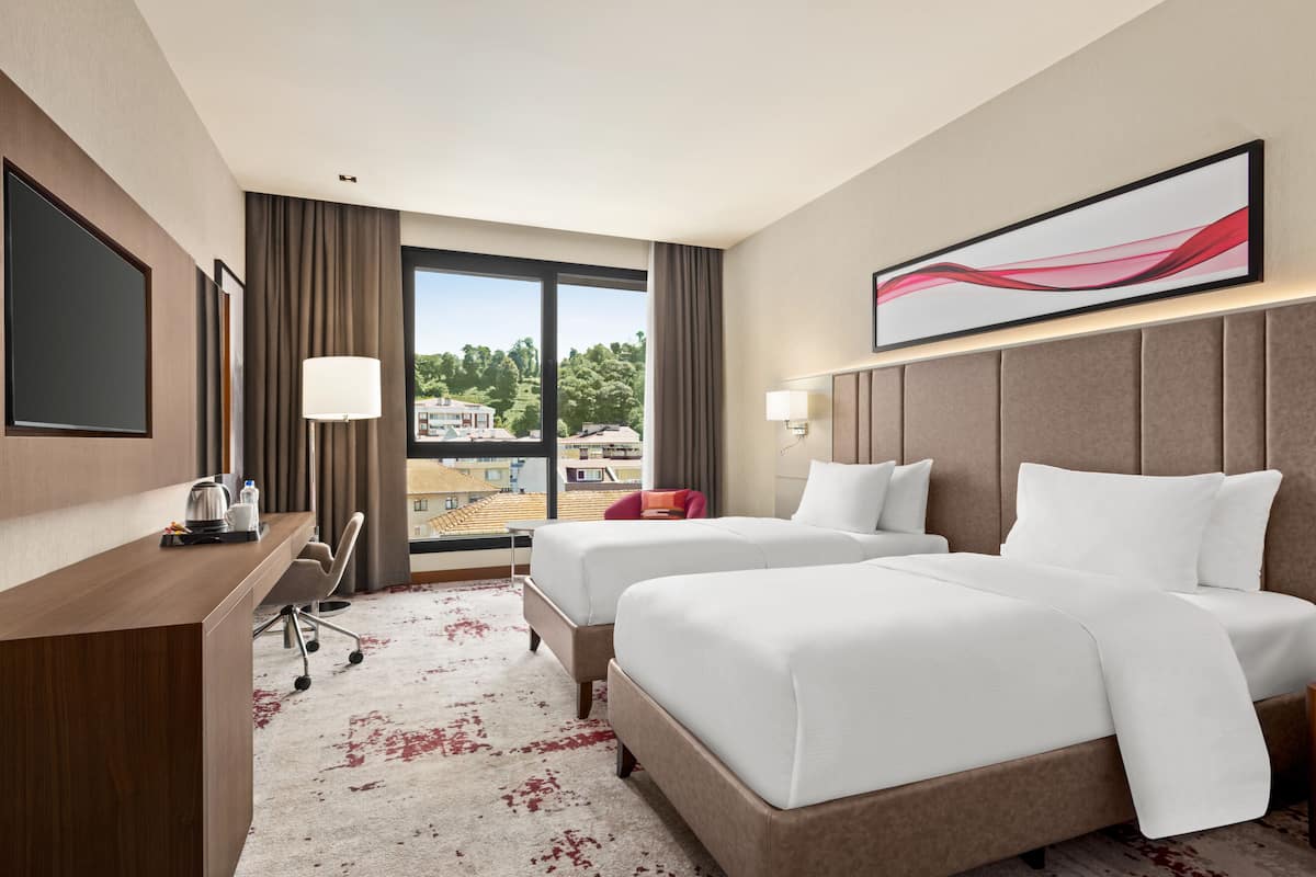 Ein Hotelzimmer mit zwei Betten, einem Schreibtisch, einem Stuhl und einem großen Fenster mit Blick auf die Stadt.