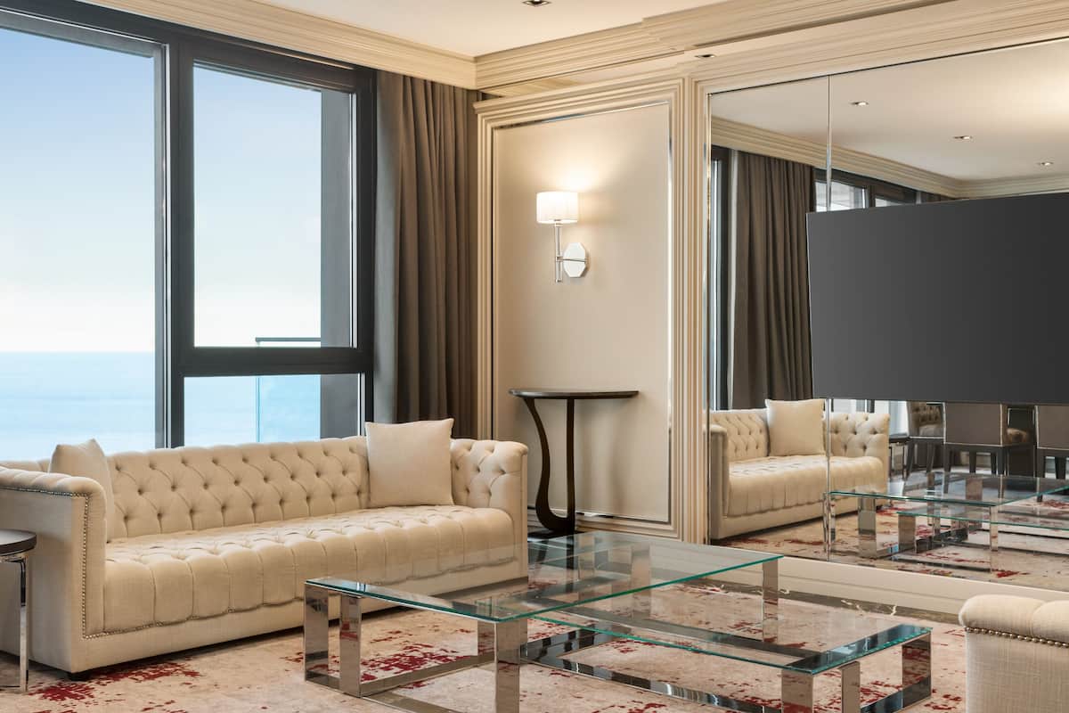 Ein modernes Hotelzimmer mit großem Fenster, einer gepolsterten Couch, einem Glastisch und einem großen Spiegel.