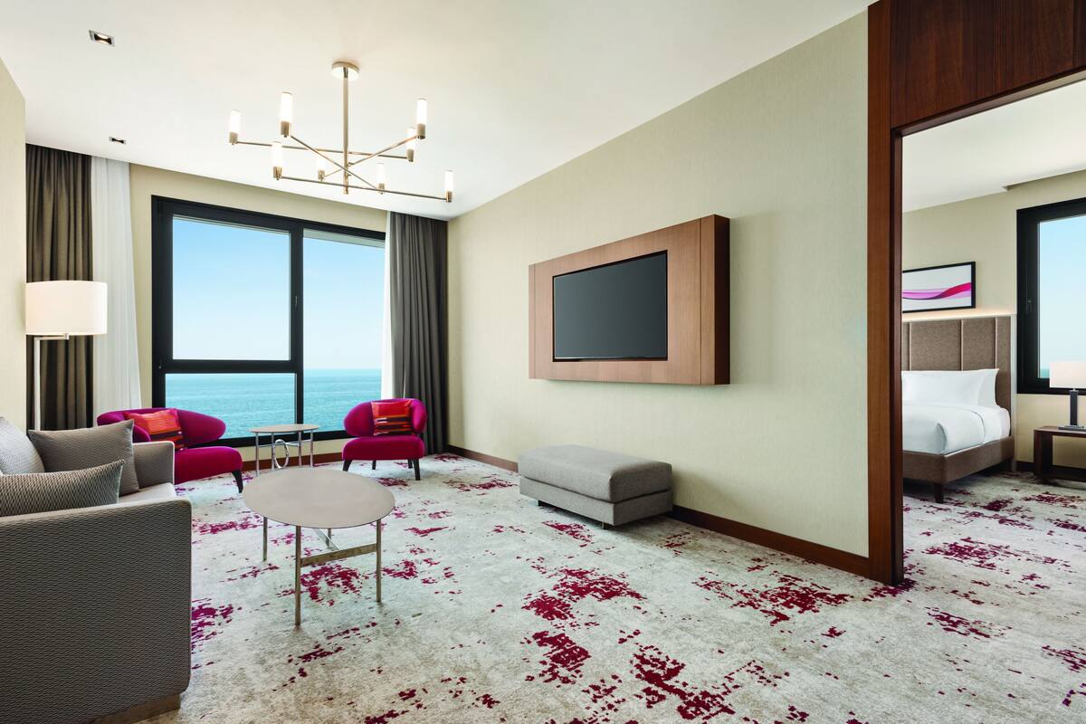 Ein Hotelzimmer mit großem Fenster, Flachbildfernseher, Sofa, Sessel, kleinem Tisch und einem Bett.