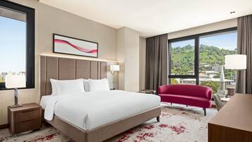 Habitación de hotel con cama, escritorio, silla, mesita de noche, lámpara, ventana con vista a un paisaje verde y una obra de arte colgada en la pared.