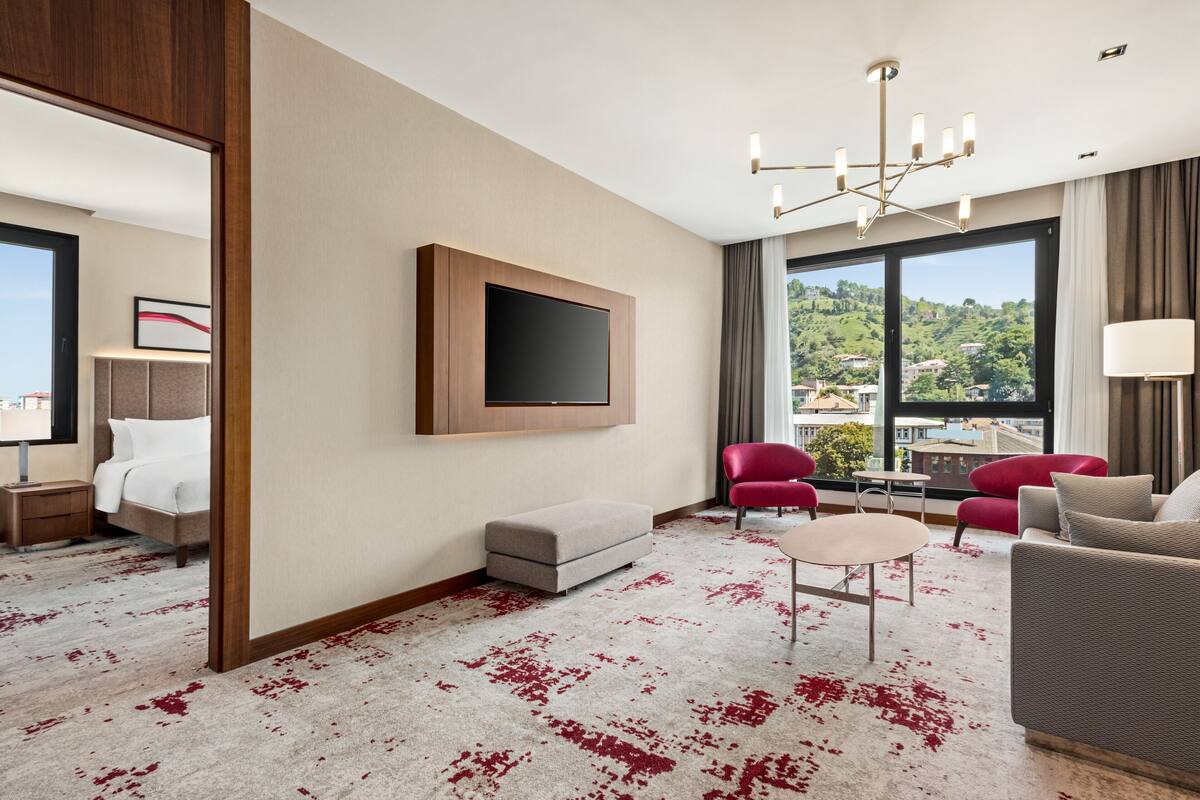 Ein modernes Hotelzimmer mit einem großen Fernseher, roten Sesseln, einer Couch, einem kleinen Tisch und Blick auf eine grüne Landschaft.