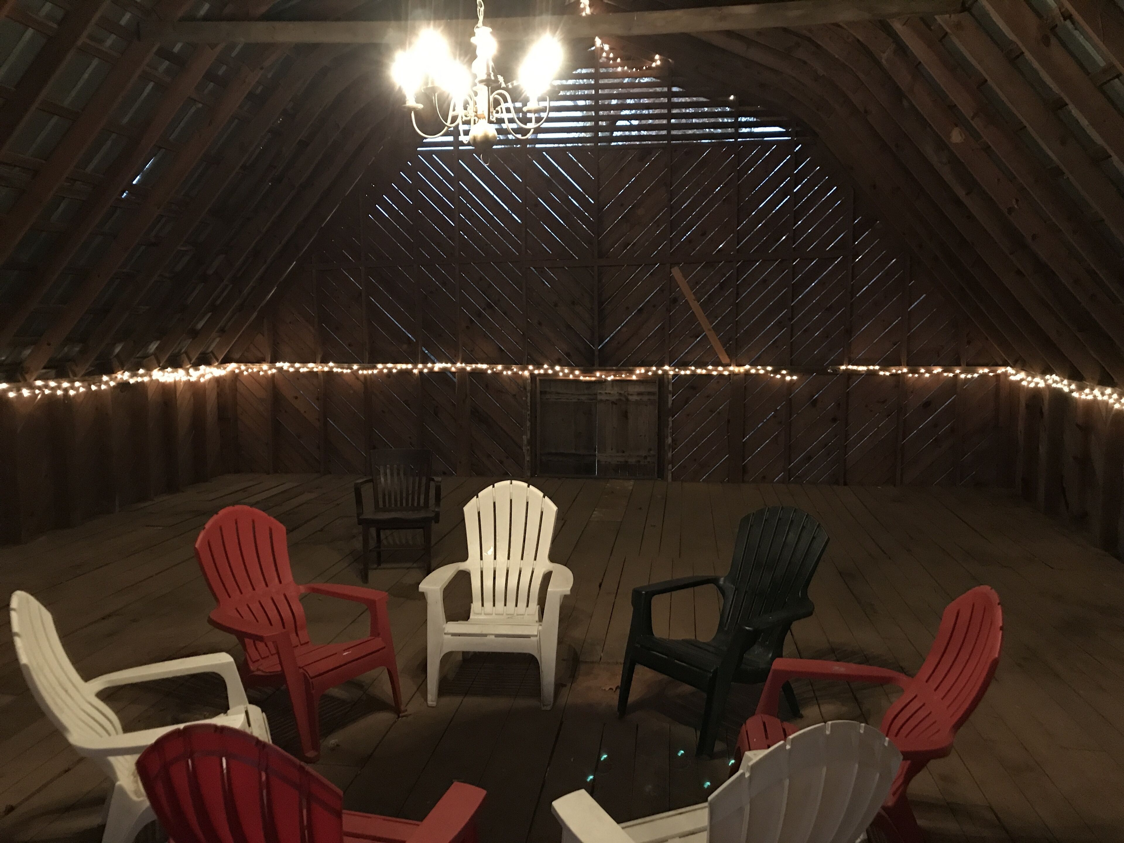 Habitación en ático con paredes de madera, un candelabro y luces colgantes. Varias sillas Adirondack están dispuestas alrededor de una pequeña mesa.