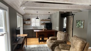 Et moderne køkken med en træø i midten, grå skabe og hængende lamper. I opholdsstuen er der en beige sofa og et spisebord med stole.