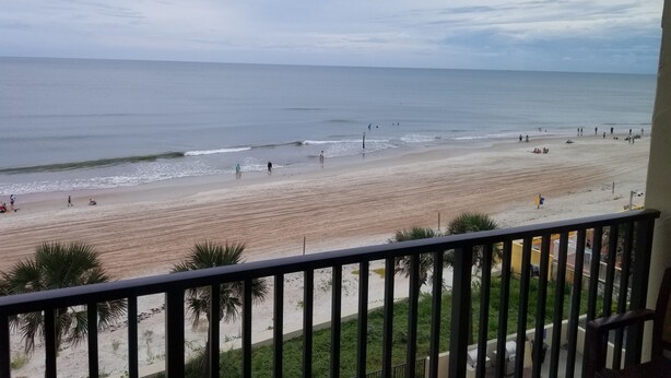 Blick auf einen Strand vom Balkon mit Geländer, Palmen und einem sandigen Strand mit Menschen.