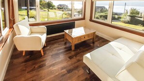 A room with a white sofa, a white armchair, a wooden coffee table, and large windows offering a view of greenery and buildings.