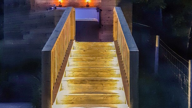 A wooden bridge with railings, leading to a building with two lit windows.