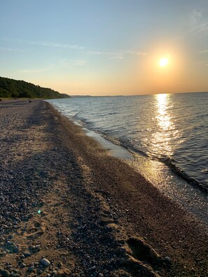 A pebbled beach with the sun reflecting on the water, surrounded by a forested hillside.