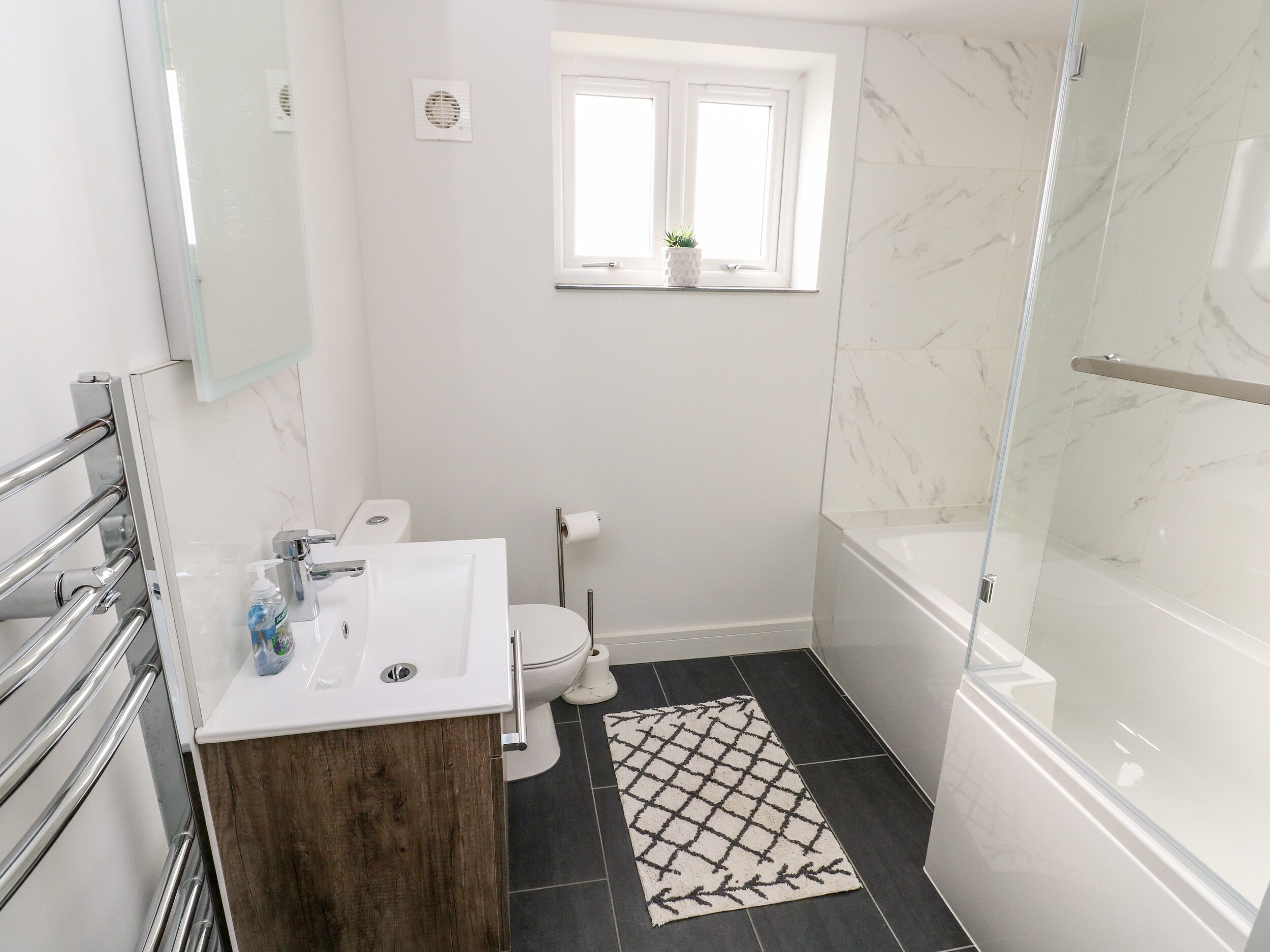 Un bagno moderno con box doccia in vetro, lavabo bianco con mobile contenitore in legno, water e una piccola finestra con una pianta.