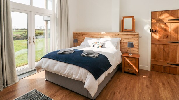 A bedroom with a large bed, wooden headboard, a nightstand, a hanging light, and a view of the outdoors through a glass door. - Glowty Brynbedw (Clynderwen)