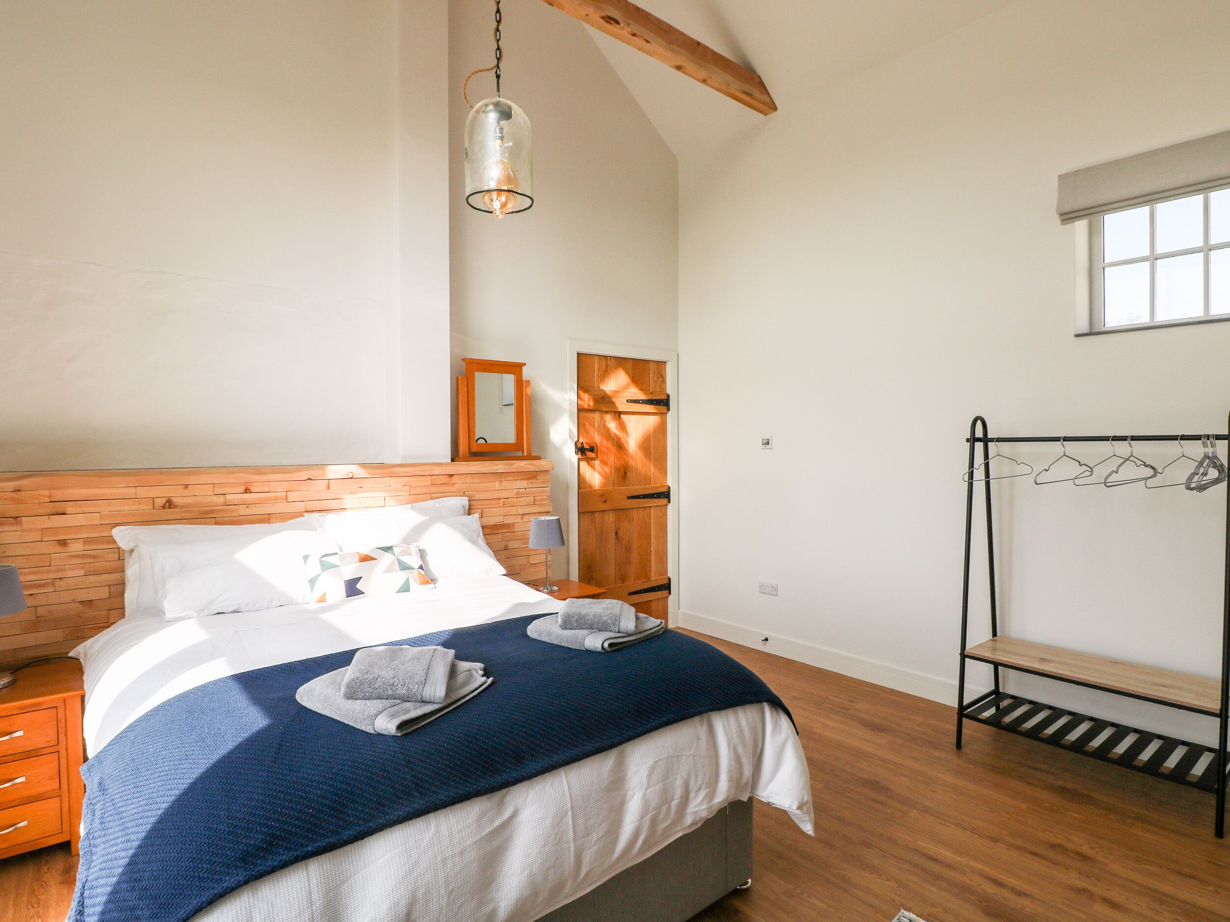 Un dormitorio con cabecera de madera, cama con manta azul, lámpara colgante, puerta de madera y un perchero con ropa.