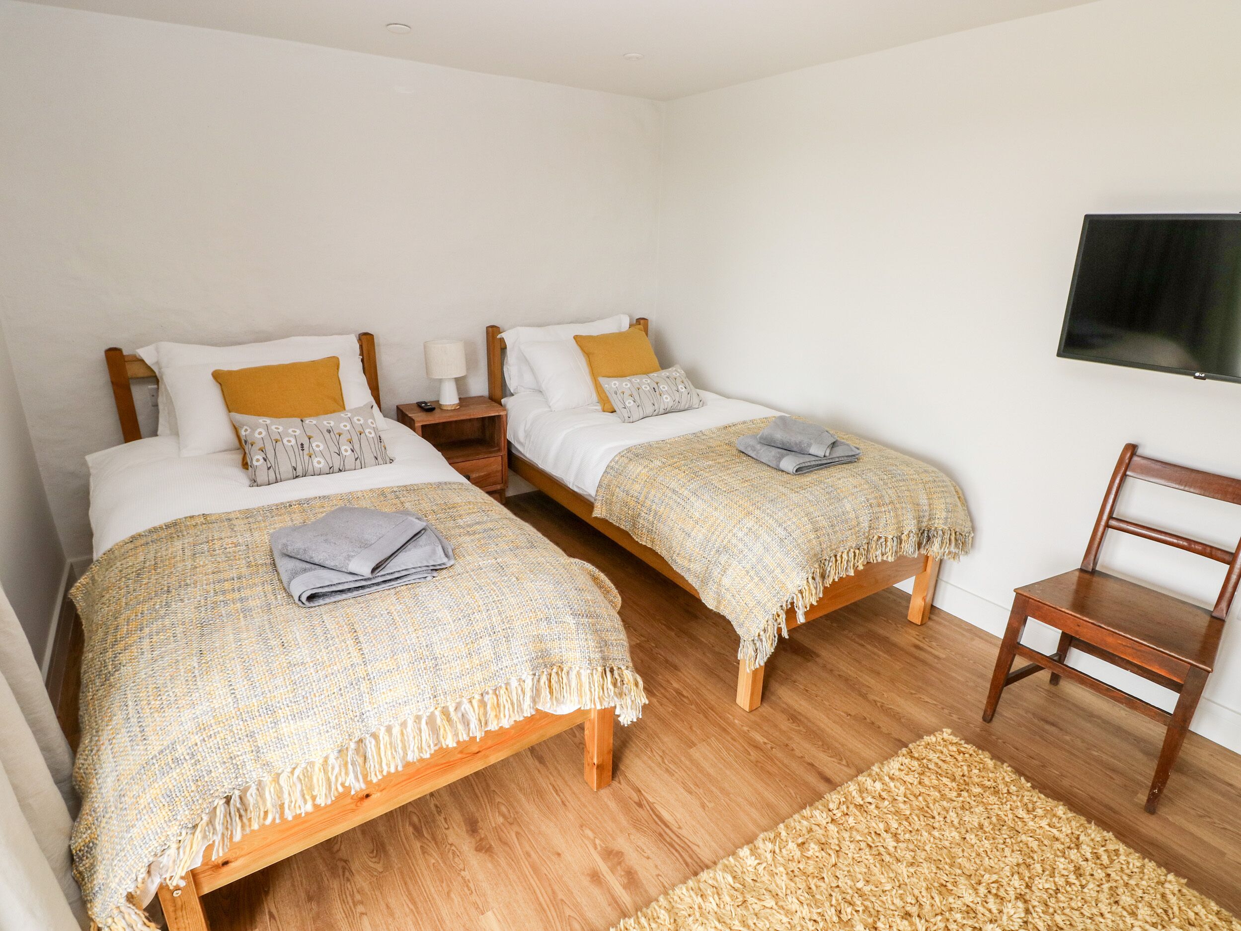 Une chambre avec deux lits en bois, chacun recouvert d’une couverture à motifs et agrémenté d’oreillers, une table de chevet en bois avec une lampe, une chaise en bois et un téléviseur fixé au mur.