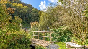 A wooden bridge over a stream, surrounded by lush greenery and a stone bench. - Wisteria Lodge - QC1677 (Preseli Hills)