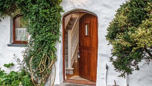 A white building with a wooden door and a window, surrounded by greenery. - Wisteria Lodge - QC1677 (Preseli Hills)
