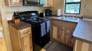 A compact kitchen with wooden cabinets, a stove, a microwave, and a sink. - Cozy Coastal Cabin #3 (Steuben)