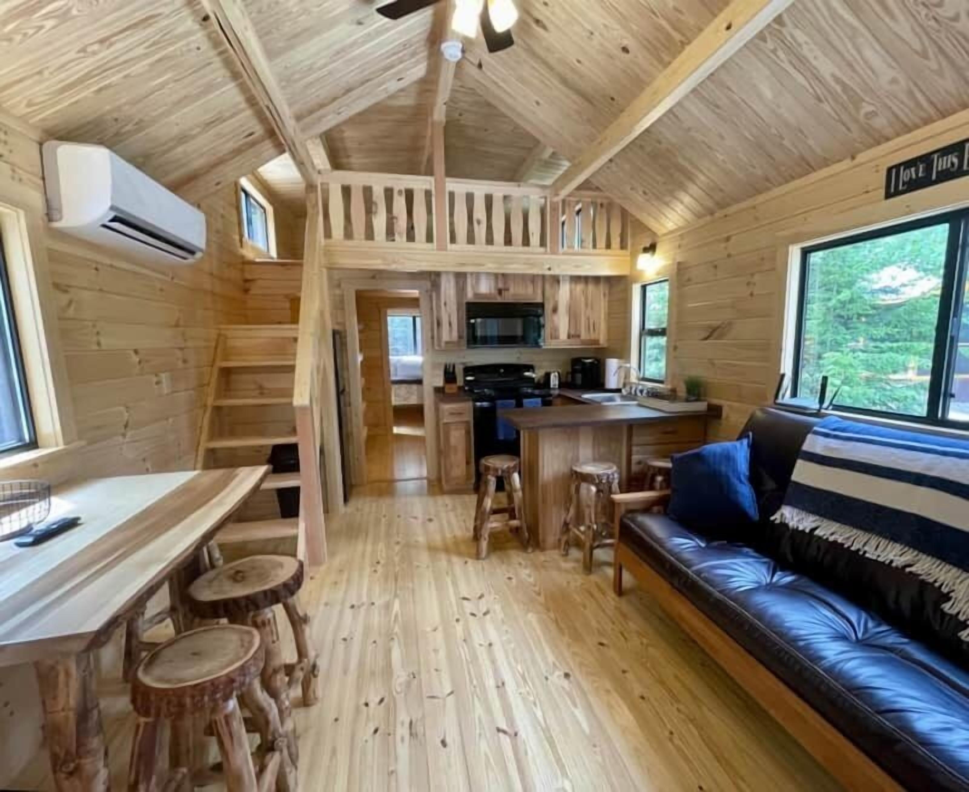 L’intérieur d’une cabane est doté d’un plancher en bois, d’un coin cuisine, d’un espace repas avec des tabourets, d’un canapé et d’un escalier menant à un niveau supérieur.