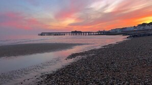 A pebbled beach with a pier extending into calm waters, buildings in the background, and a sunset sky. - Number 46 (Hastings)