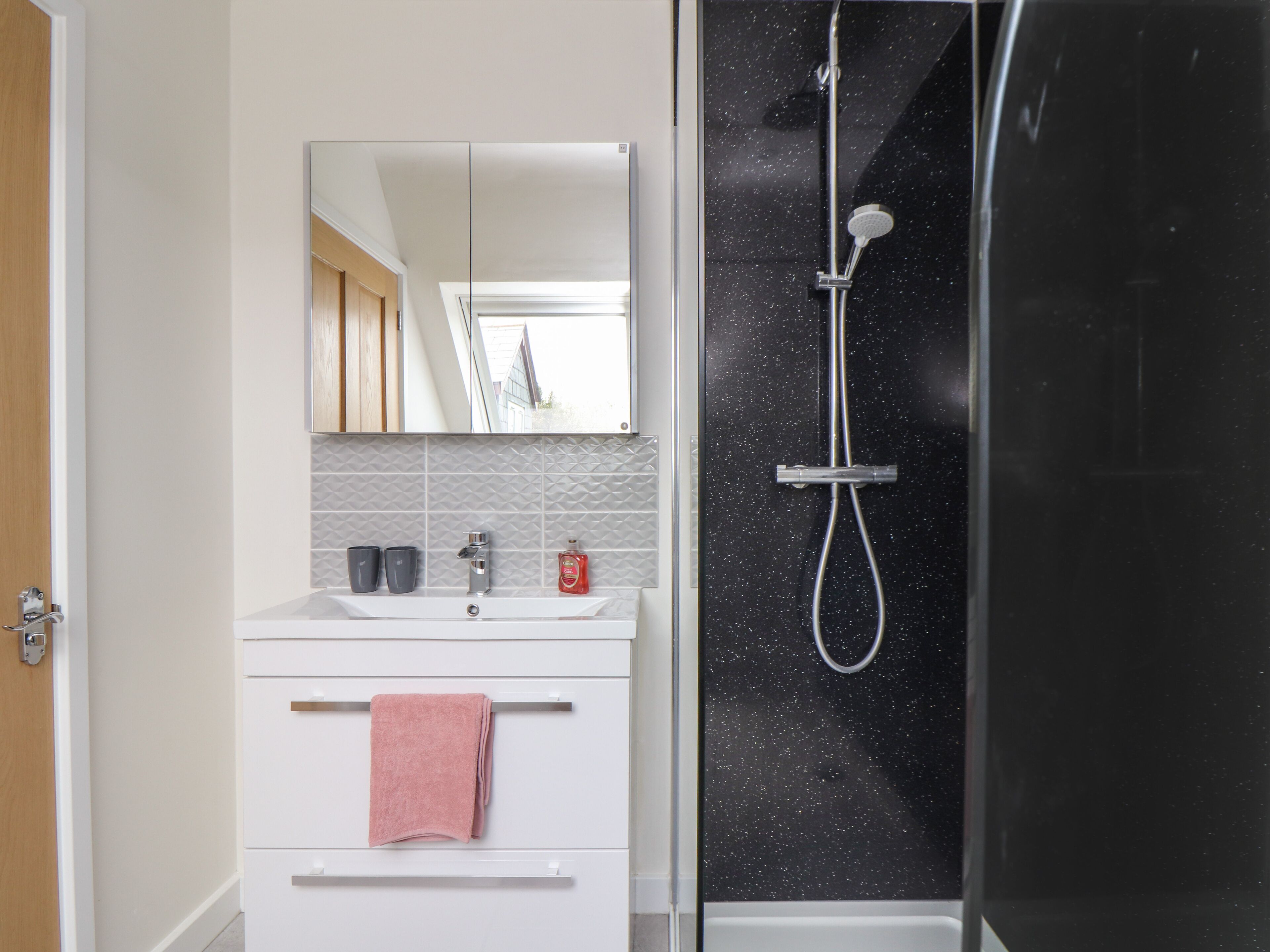 Une salle de bain avec un meuble-lavabo blanc, un miroir, un évier, une serviette et une douche dont le mur est noir et parsemé de petites taches.
