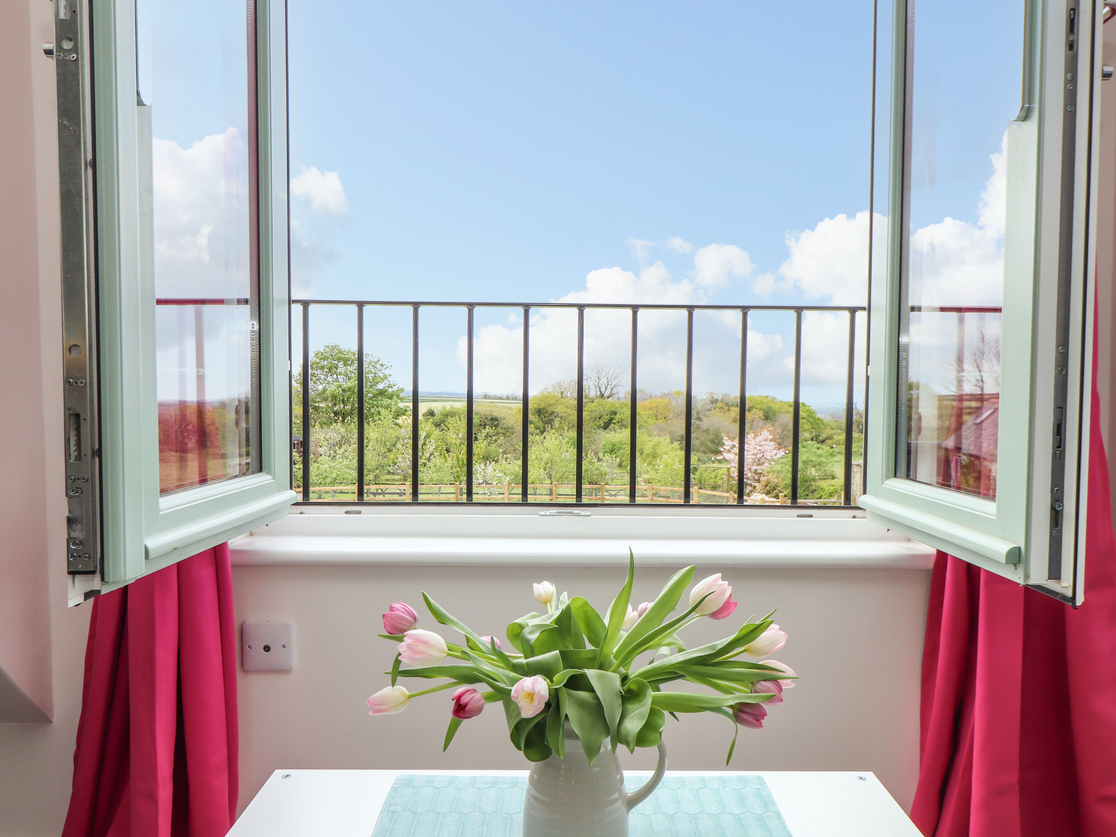 A room with a window overlooking a balcony with a metal railing, a vase with flowers on a table, and red curtains.