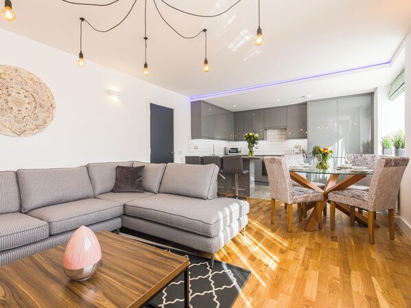 A modern living room with a sectional sofa, a glass dining table, and a kitchen in the background.