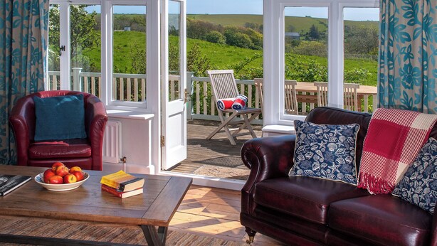 A cozy room with a leather sofa, a wooden coffee table with a bowl of apples and books, and a view of a green landscape through large windows. - Manorbier Boat House (Manorbier)