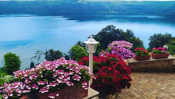 Vista de un lago con montañas al fondo, rodeado de exuberante vegetación y parterres de flores.