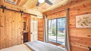 Ein Schlafzimmer einer Holzhütte mit einem Bett, einem Deckenventilator, einer Schiebetür und einem großen Fenster.
