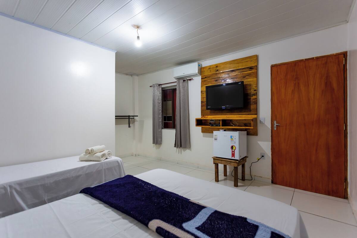 A hotel room with two beds, a television mounted on a wooden panel, a small refrigerator, and a wooden wardrobe.