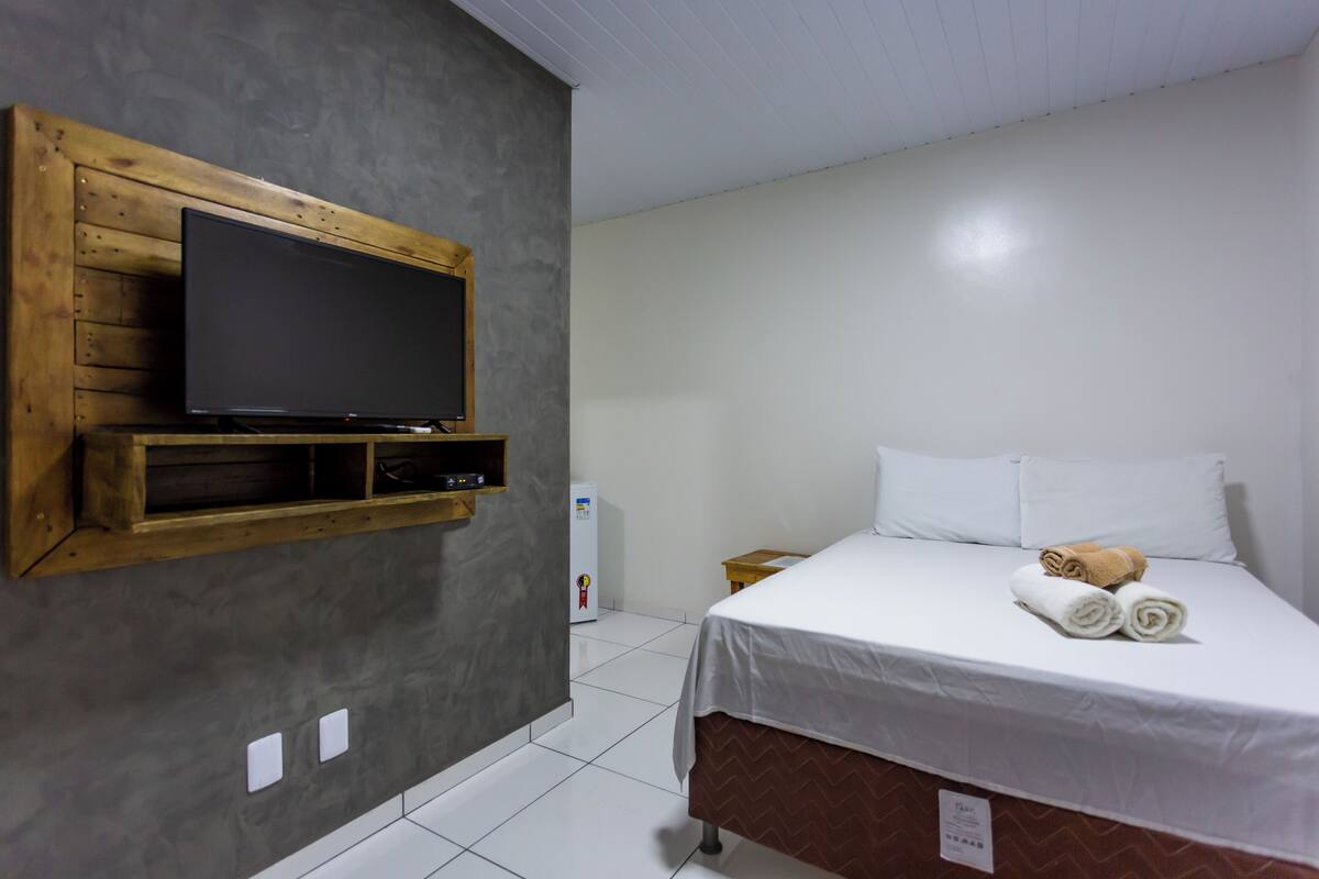A hotel room with a bed, a TV mounted on a wooden panel, and a small bedside table.