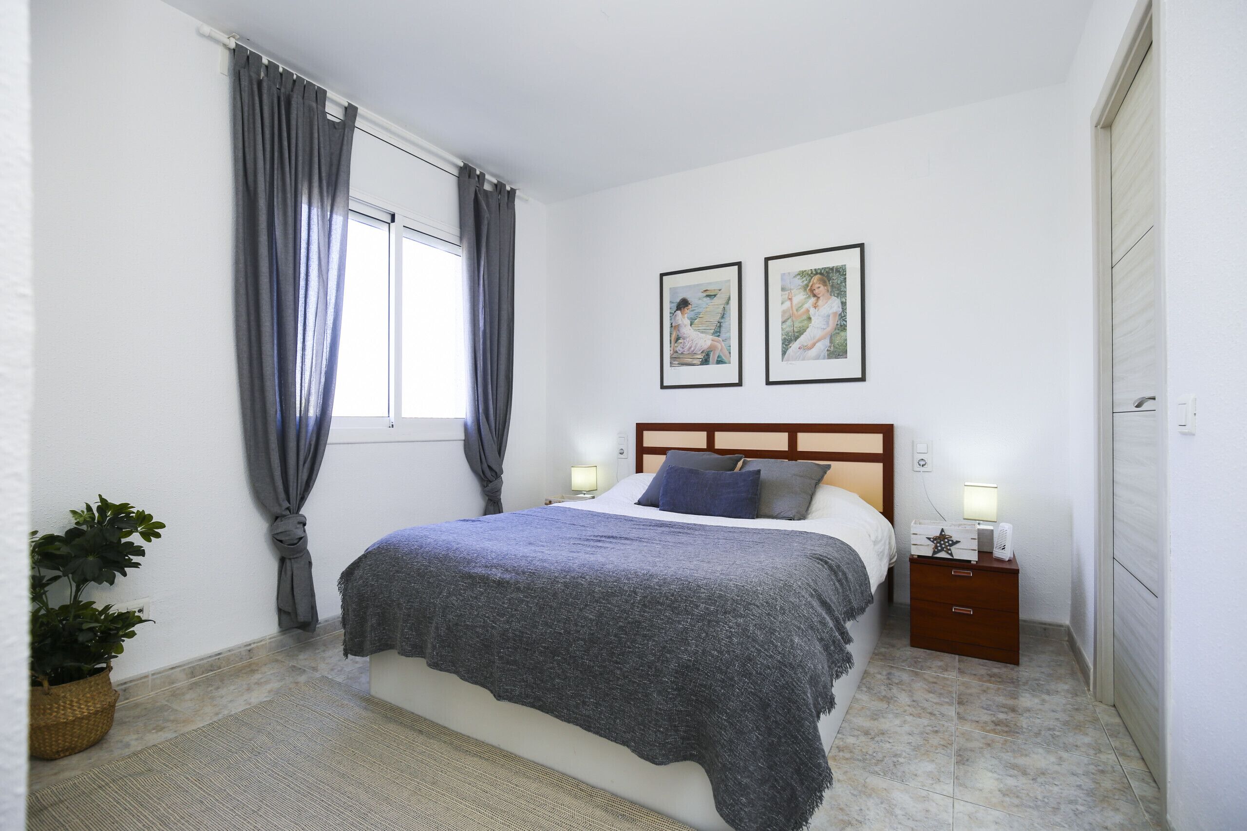 Une chambre à coucher comprenant un lit, deux tables de chevet, une petite table, une plante en pot et deux tableaux encadrés accrochés au mur.