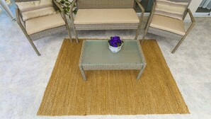Een rieten zithoek met beige kussens, een geweven tafel en een geel vloerkleed.