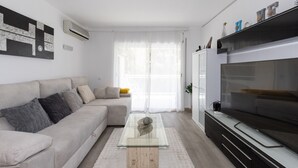 Een moderne woonkamer met een bank, een glazen salontafel, een flatscreen televisie en een groot raam met dunne gordijnen.