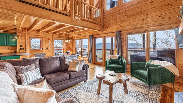 A spacious log cabin interior with a large living area, a dining area, and large windows offering views of the outdoors.