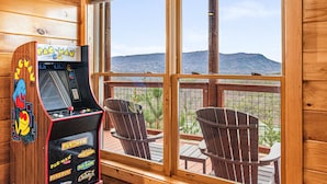 A wooden cabin with a Pac-Man arcade machine, two Adirondack chairs, and a view of mountains through large windows.