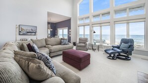A spacious living room with a large window overlooking the sea, a comfortable sofa set, a recliner, and a small table. - Exquisite Private Lake Front Family Home (Manistee)