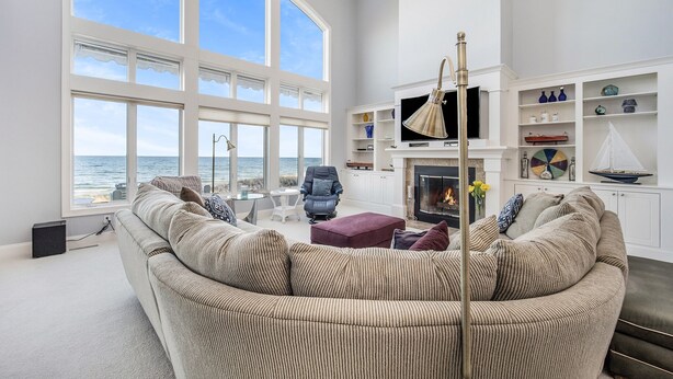 A spacious living room with a large window overlooking the sea, a comfortable sofa, a fireplace, and a well-organized bookshelf. - Exquisite Private Lake Front Family Home (Manistee)