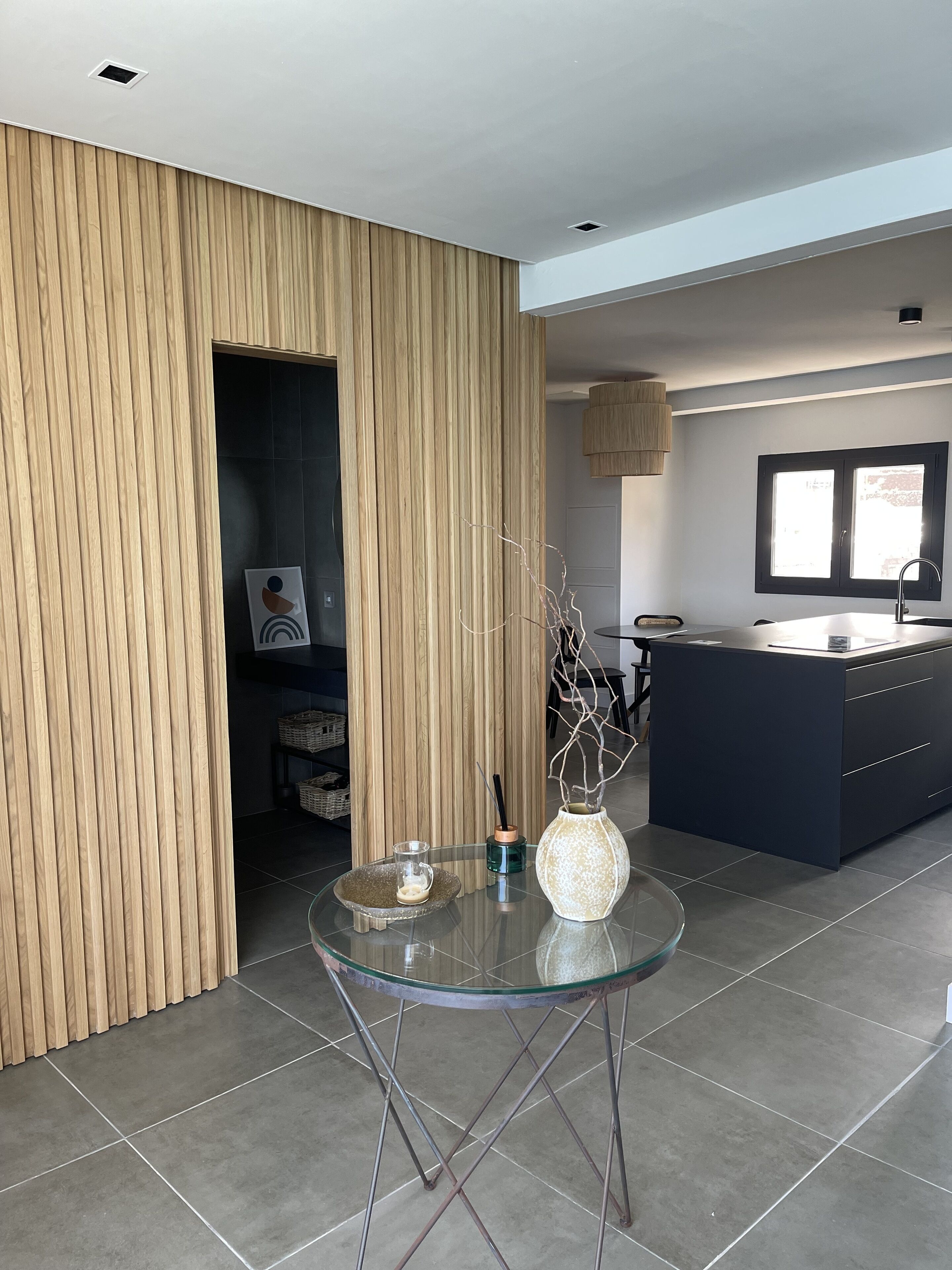 Une cuisine moderne avec un mur d’accent en bois, une table en verre agrémentée d’un vase et un îlot central foncé doté d’un évier.