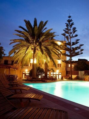 Zona de piscina con sillones de descanso y un edificio de varios pisos con balcones.