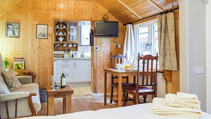 A cozy room with wooden walls and ceiling, a dining table with chairs, a TV mounted on the wall, and a kitchen area with cabinets and a refrigerator. - Woodcarver's Cottage (Spring Village)