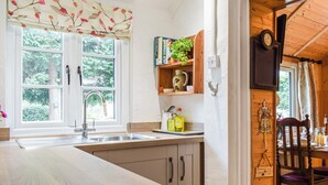 A kitchen with a wooden countertop, a sink, and a window with a view of greenery. There is a shelf with decorative items and a clock on the wall. - Woodcarver's Cottage (Spring Village)