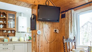 A wooden-paneled kitchen with white cabinets, a television mounted on the wall, and a window with curtains. - Woodcarver's Cottage (Spring Village)