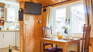 A wooden interior with a dining table set for breakfast, a television mounted on the wall, and a window with curtains. - Woodcarver's Cottage (Spring Village)