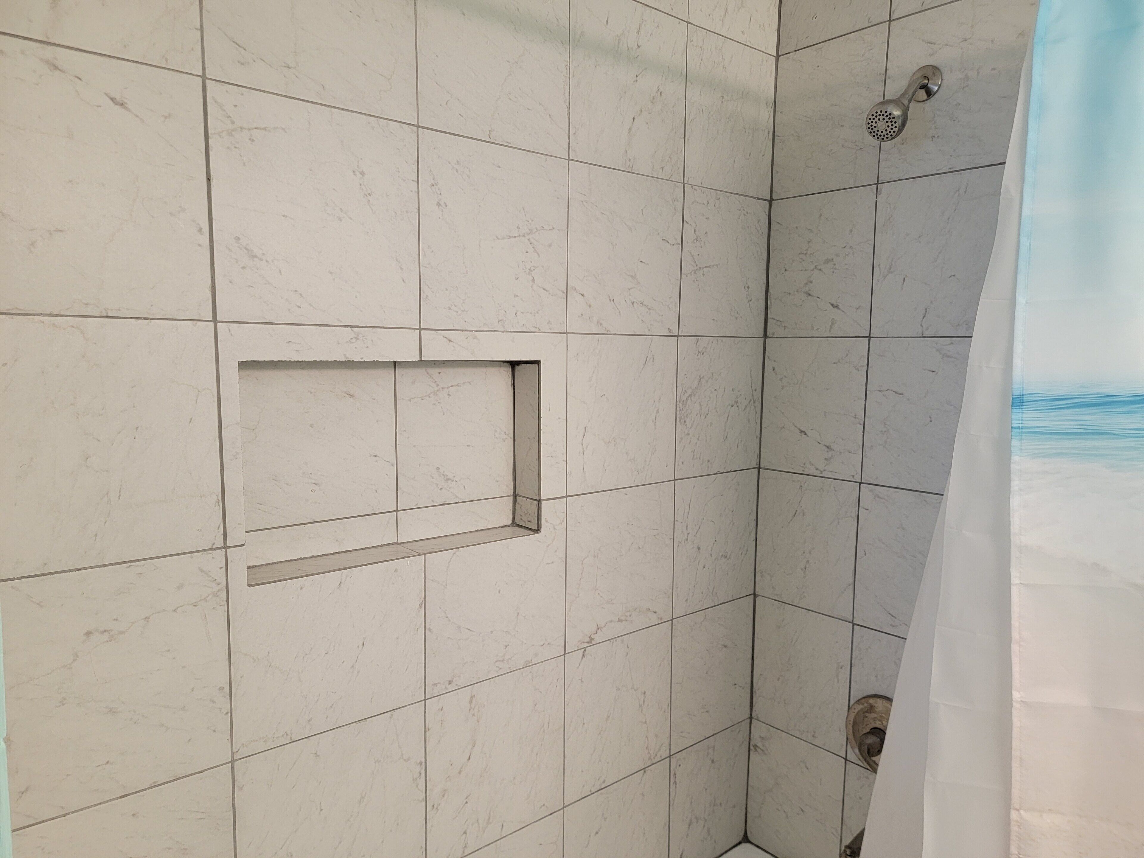 Un baño con baldosas de mármol blanco, una ducha con cabezal de ducha manual y un estante rectangular pequeño.