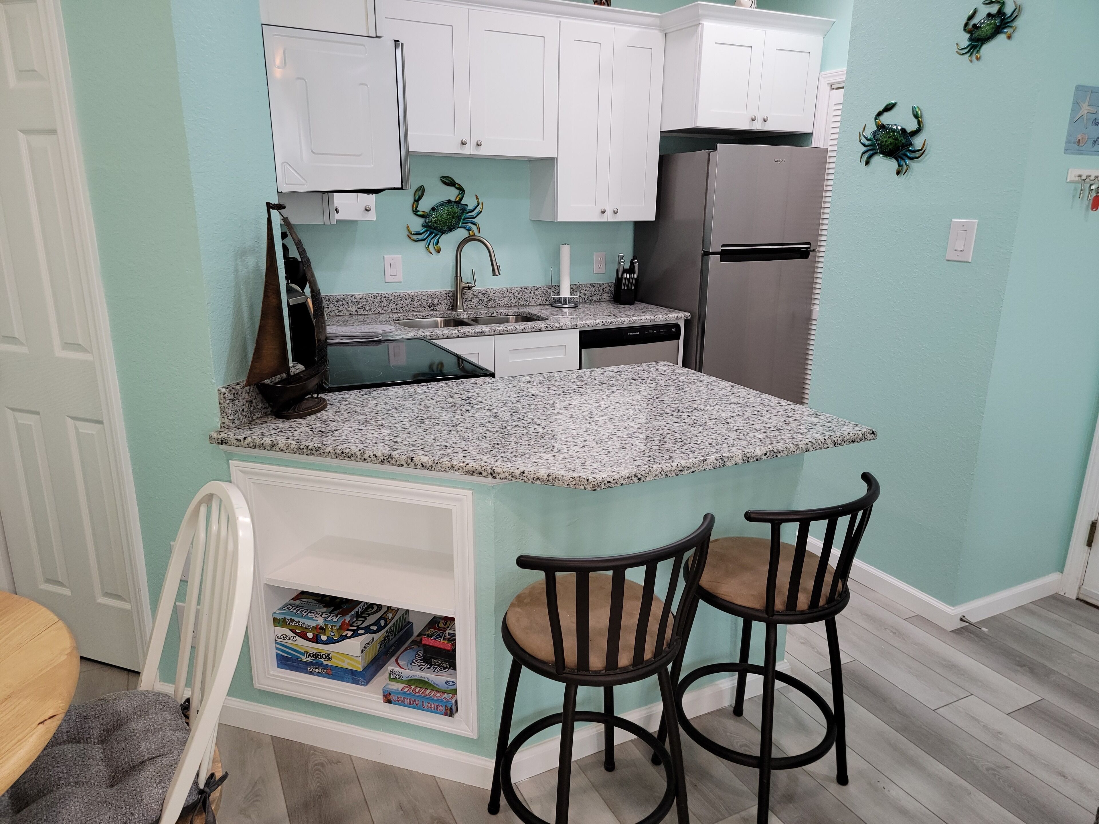 A compact kitchen with white cabinets, a stainless steel refrigerator, granite countertops, and two bar stools.