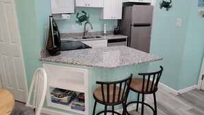 Cocina compacta con armarios blancos, refrigerador de acero inoxidable, encimeras de granito y dos taburetes de bar.